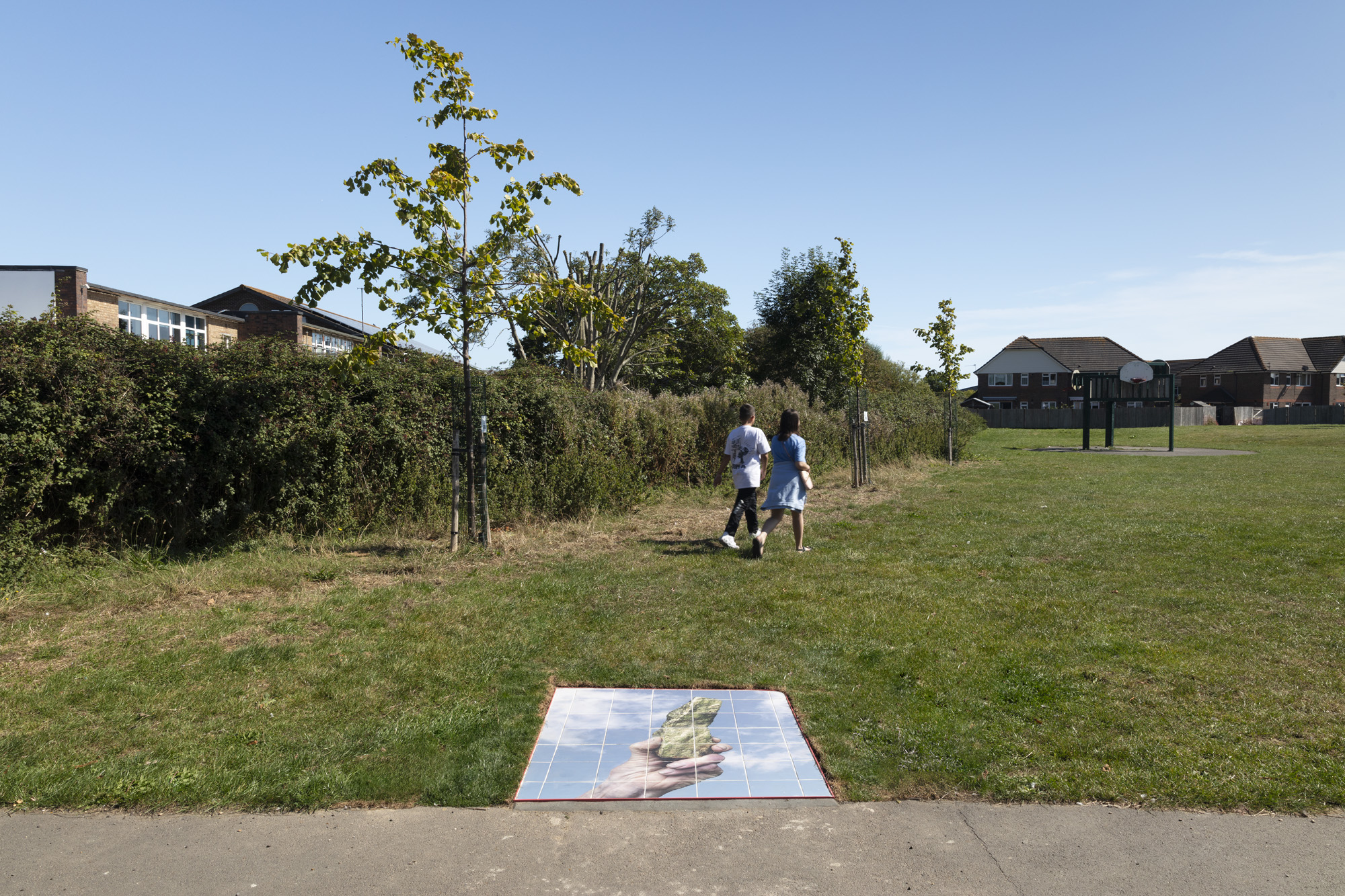 Tiles printed with a photograph of a hand holding a bronzed object against a cloudy sky are embedded into the grass at Langney Playing Fields